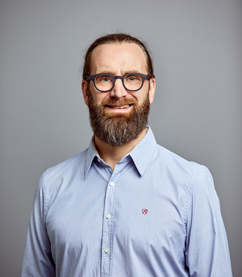 Mann mit Vollbart und Brille vor grauem Hintergrund lächelt in die Kamera